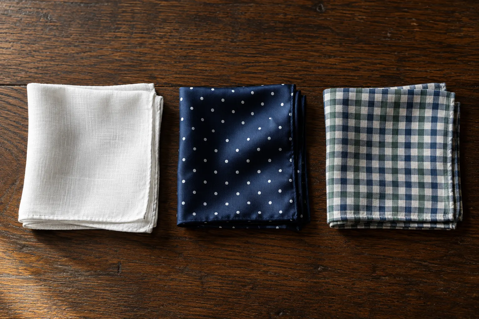 White linen, navy polka-dot silk and blue and green check cotton pocket squares laid side by side on a dark oak table